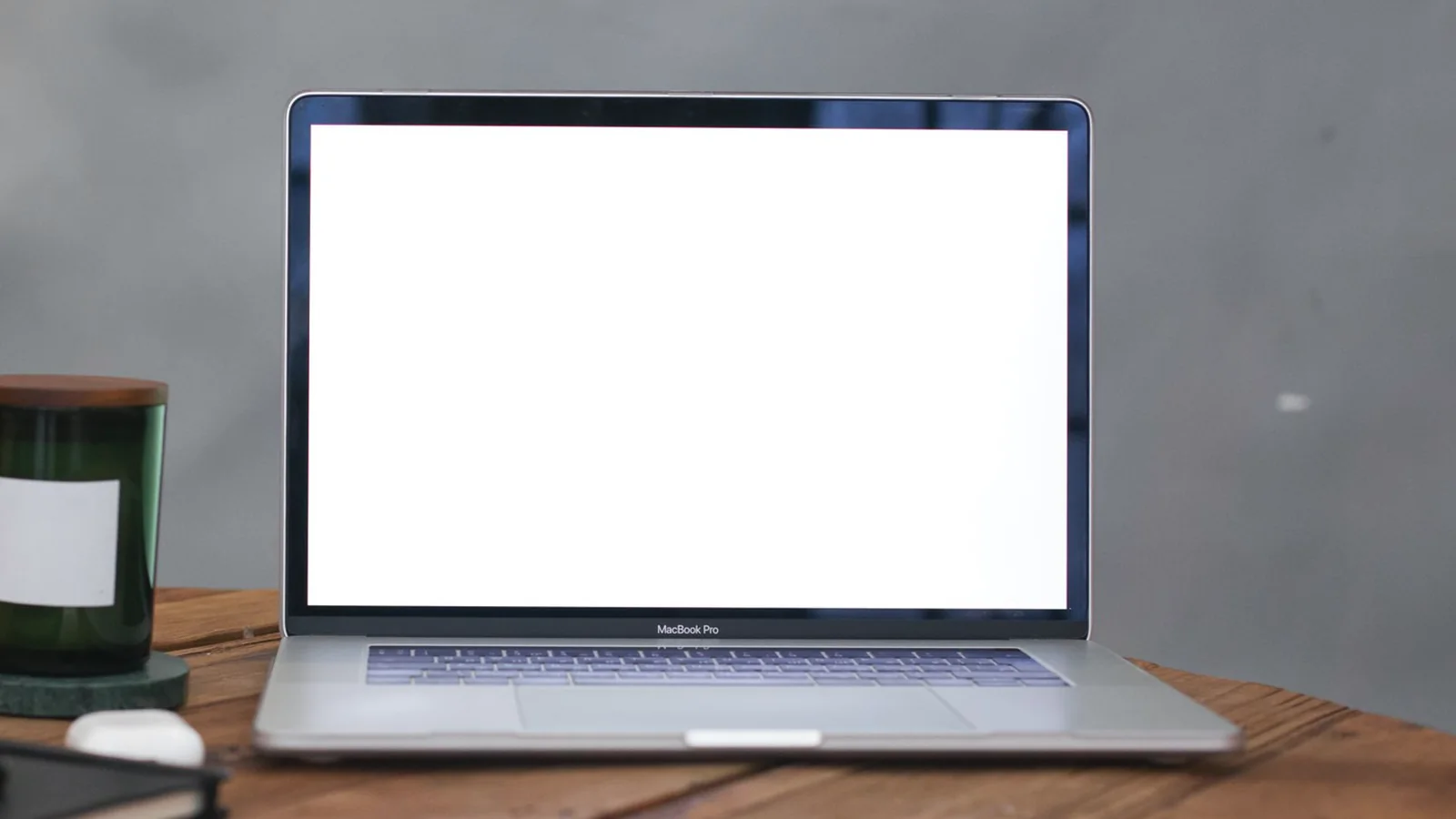 Aufgeklappter Laptop und ein Glasgefäß auf einem Holztisch vor einer grauen Wand.