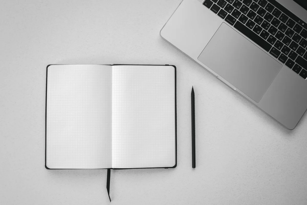 Aufgeschlagenes leeres Notizbuch mit Stift neben einem Laptop auf hellem Tisch.